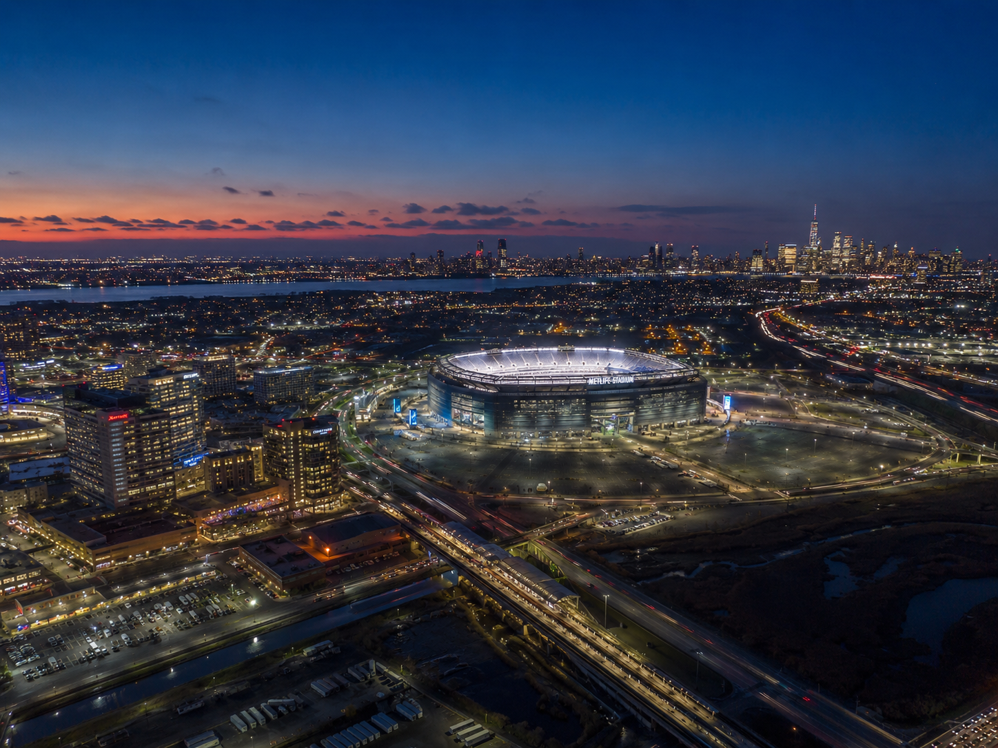 Aerial view of MetLife Stadium, nearby New Jersey hotels, and distant New York City skyline at night.
